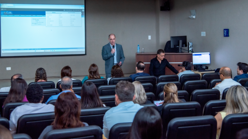 A speaker presents to an audience in a conference room, with a projector displaying information on a screen.