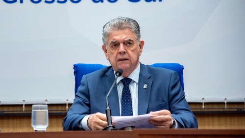 A man in a suit speaks at a podium, holding papers, with a glass of water nearby. A blue backdrop is visible behind him.