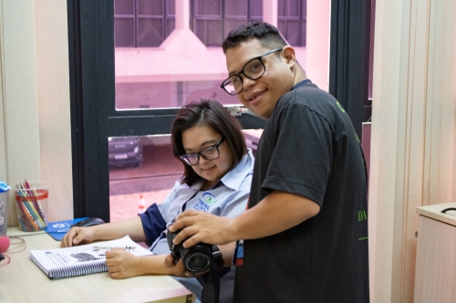 A man and a woman are in an office setting. The woman is seated at a desk, while the man stands beside her, holding a camera.