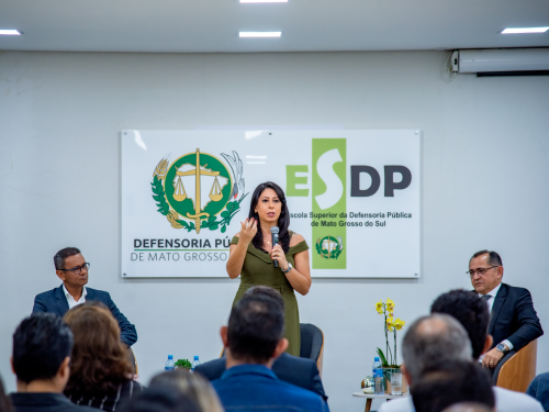 A woman speaks at a podium in front of a banner for the Public Defender's Office of Mato Grosso do Sul, with two men seated nearby.