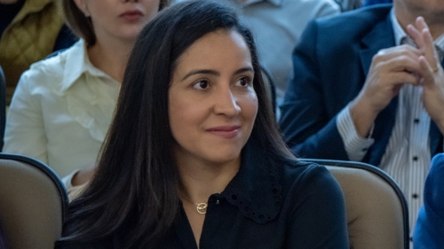 TCE-MS realiza prévia de curso sobre o sistema e-Sfinge com servidores do Governo do Estado A woman with long dark hair is seated, smiling slightly, in a crowd during an event. She wears a black top and appears engaged.
