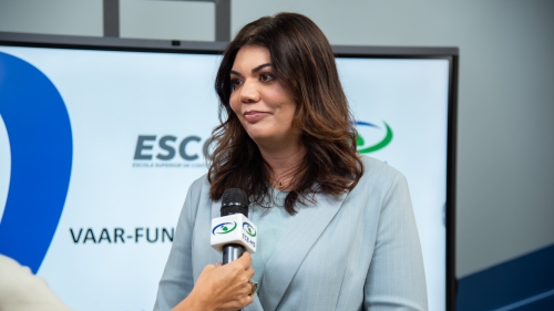TCE-MS faz live para orientar municípios sobre recursos do VAAR A woman with long dark hair is being interviewed, wearing a light blue blazer, in front of a screen displaying logos.