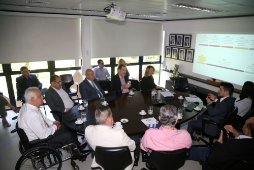 A group of people is seated around a conference table, engaged in discussion. A presentation is displayed on a screen.