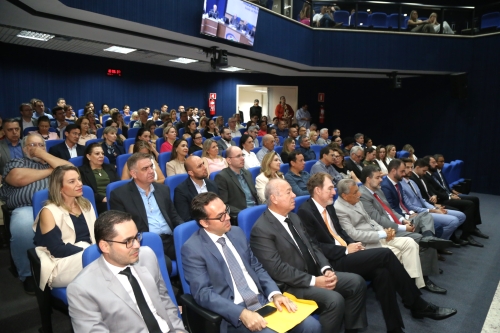 A large audience seated in a conference room, with a mix of men and women in formal attire, facing a speaker at the front.