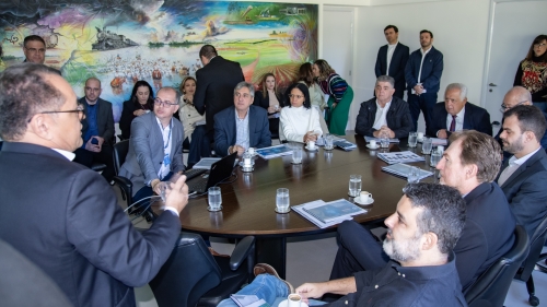 A group of people in business attire is seated around a conference table, engaged in discussion. A colorful mural is visible in the background.