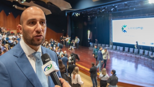 TCE-MS participa do 1º Congresso Estadual de Licitações e Compras Públicas de MS A man in a light blue suit speaks into a microphone at an event with an audience and a stage in the background.