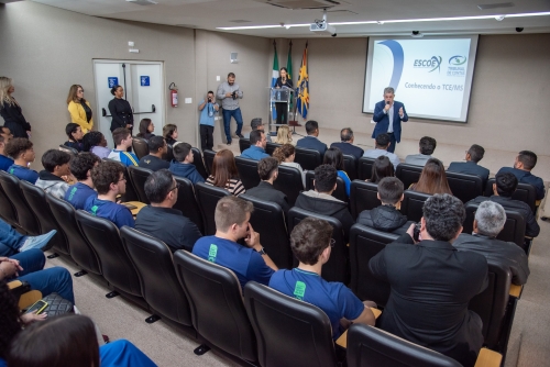 Comitiva de estudantes e servidores de Chapadão do Sul conhecem o TCE-MS A conference in a lecture hall with an audience seated, listening to a speaker at the front. Flags are displayed in the background.