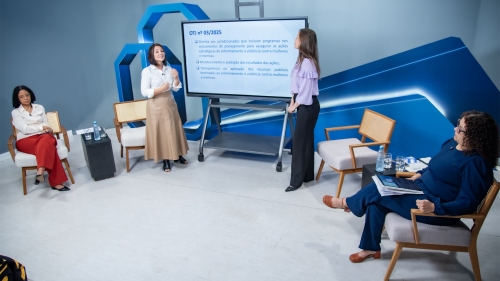 A presentation scene with four women. One is speaking while another presents on a screen, and two others are seated, observing.