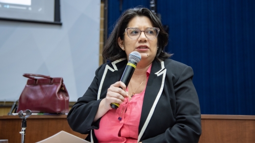 TCE-MS ouve gestoras municipais e propõe ação conjunta para fortalecer políticas públicas às mulheres A woman with glasses speaks into a microphone, wearing a black blazer over a pink shirt, seated at a table with a bag in the background.