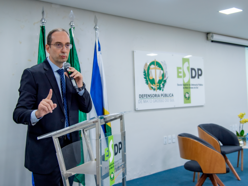 A man in a suit speaks at a podium with flags in the background, in a formal setting. A sign reads "DEFENSORIA PUBLICA DE MATO GROSSO DO SUL."