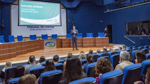 A speaker addresses an audience in a formal setting, with a presentation screen displaying "DIÁLOGO INTERNO" in a blue-themed auditorium.