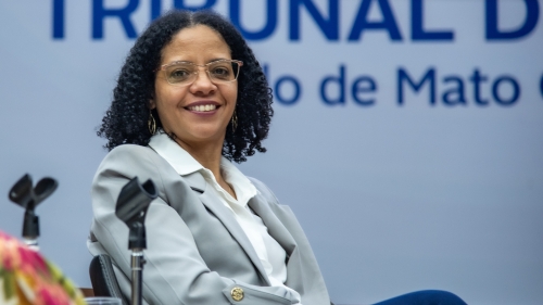 TCE-MS ouve gestoras municipais e propõe ação conjunta para fortalecer políticas públicas às mulheres A woman with curly hair and glasses smiles while seated, wearing a light gray blazer over a white shirt, against a backdrop with text.