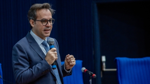 A man in a suit speaks into a microphone, gesturing with his hands. Blue chairs are visible in the background.