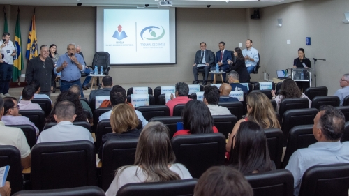 A conference or meeting in a large room with an audience seated. Speakers are at the front, with flags and a presentation screen visible.
