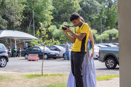 A young man in a yellow shirt is holding a camera, while a woman in a long dress stands nearby. Cars are parked in the background.