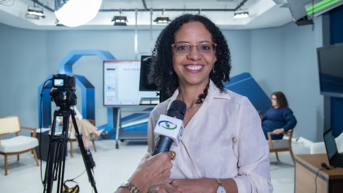 A woman with curly hair smiles while holding a microphone in a studio setting, with cameras and a screen in the background.