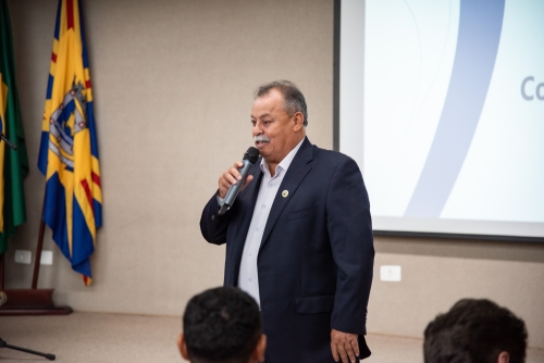 Comitiva de estudantes e servidores de Chapadão do Sul conhecem o TCE-MS A man in a suit speaks into a microphone, with flags in the background. He appears to be addressing an audience.
