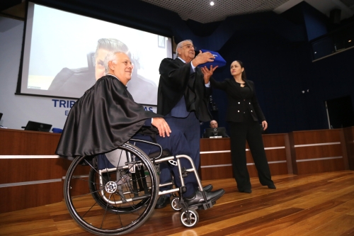 A man in a wheelchair is receiving an award or recognition, while another man gestures expressively. A woman stands nearby, observing.