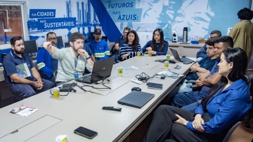 TCE-MS entrega manual de operação do equipamento ZFG 3000 à Águas Guariroba A group of people is seated around a conference table, engaged in a discussion. A presenter gestures towards a laptop.