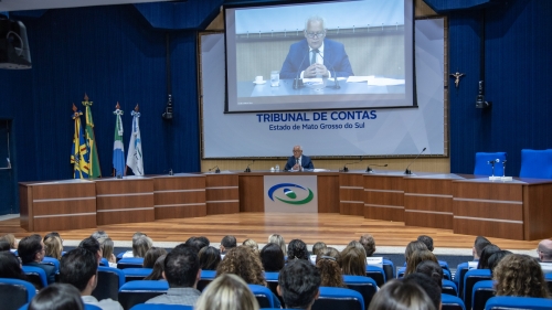 TCE-MS lança plataforma de Inteligência Artificial Generativa e guia de uso ético da tecnologia A conference room with a speaker at a podium, facing an audience. Flags are displayed, and a large screen shows the speaker.