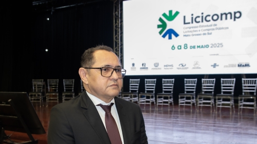 TCE-MS participa do 1º Congresso Estadual de Licitações e Compras Públicas de MS A man in a suit sits in front of a large screen displaying event details for "Licicomp" in a conference setting.