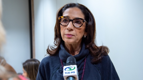 A woman with long, dark hair and glasses speaks into a microphone, wearing a dark sweater in a well-lit room.