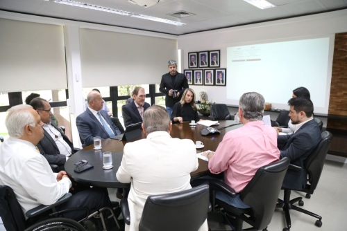 A group of professionals in a conference room, seated around a circular table, engaged in discussion with a presentation on a screen.