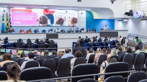A conference or meeting in a large room with a panel of speakers, audience seated, and a presentation displayed on a screen.
