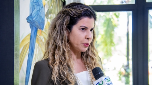 MPMS e Defensoria pedem ajuda ao TCE-MS para solucionar problema de contratação de professores de apoio A woman with curly hair speaks into a microphone, set against a backdrop of a colorful parrot painting.