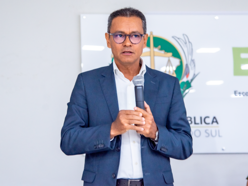 A man in a suit stands speaking into a microphone, with a backdrop featuring a coat of arms and text.