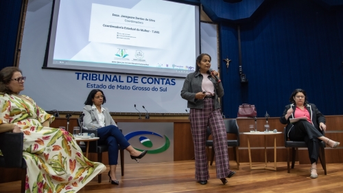 TCE-MS ouve gestoras municipais e propõe ação conjunta para fortalecer políticas públicas às mulheres A woman speaks at a podium during a conference, with three seated panelists in a formal setting. A presentation is displayed behind her.