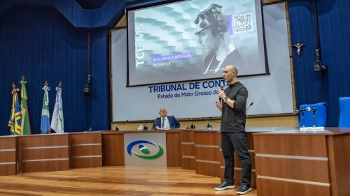 TCE-MS lança plataforma de Inteligência Artificial Generativa e guia de uso ético da tecnologia A speaker presents on stage in front of a large screen at a conference, with flags displayed in the background.