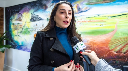 TCE-MS lança projeto para apoiar municípios na implementação da LGPD A woman speaks into a microphone in front of a colorful mural depicting landscapes. She wears a black coat and a blue turtleneck.