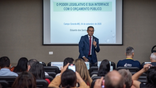 A speaker presents at a conference, discussing legislative power and public budgeting, with an audience engaged in the background.