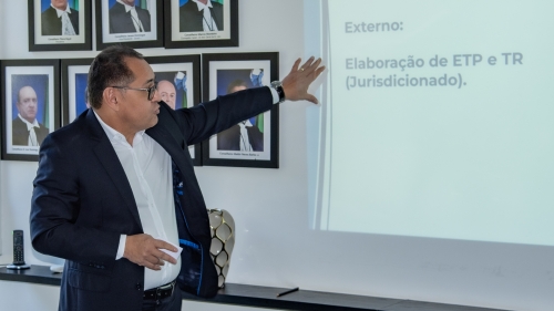 A man in a suit gestures while presenting information on a screen, with framed portraits in the background.