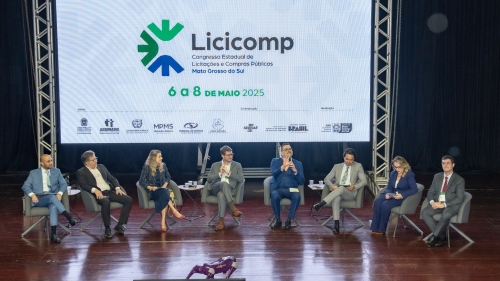 TCE-MS participa do 1º Congresso Estadual de Licitações e Compras Públicas de MS A panel discussion featuring seven speakers on stage, with a large screen displaying the event logo and details in the background.