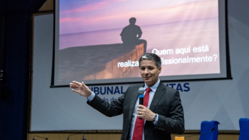 Auditoria Baseada em Riscos é tema de capacitação para auditores e técnicos do TCE-MS A man in a suit speaks at a podium, gesturing towards a presentation screen with a sunset image and text in Portuguese.