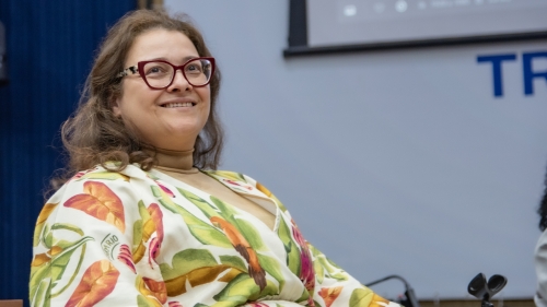 TCE-MS ouve gestoras municipais e propõe ação conjunta para fortalecer políticas públicas às mulheres A smiling woman with glasses and curly hair, wearing a colorful leaf-patterned shawl, seated in a conference setting.
