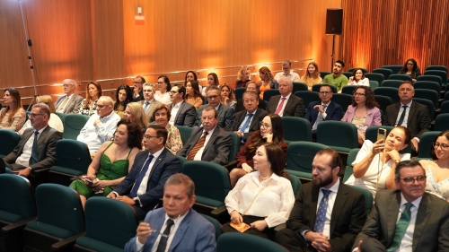 A large audience seated in a modern auditorium, dressed in formal attire, with a warm wooden backdrop and soft lighting.