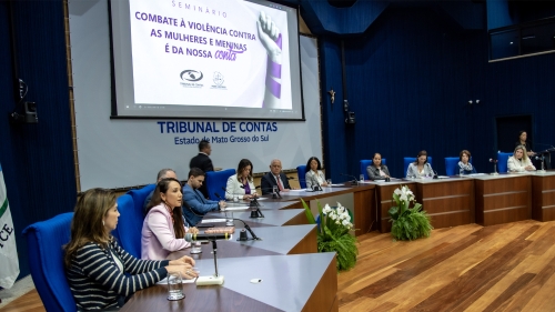 TCE-MS ouve gestoras municipais e propõe ação conjunta para fortalecer políticas públicas às mulheres A seminar on combating violence against women and girls is taking place at a conference table, with multiple speakers and an audience present.