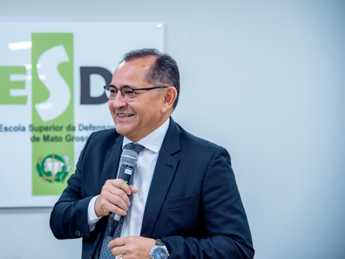 A man in a suit smiles while holding a microphone, standing in front of a sign for the Escola Superior da Defesa de Mato Grosso.