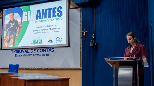 A woman stands at a podium in a conference room, with a presentation screen displaying "GIGANTES do Controle Externo."