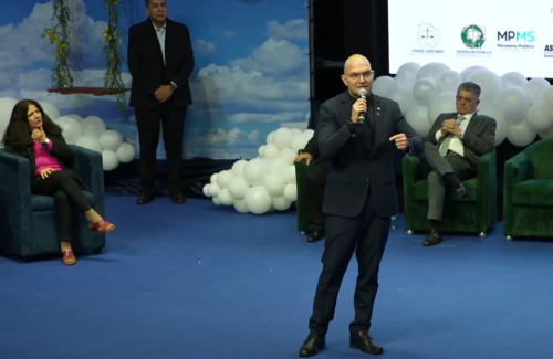 A speaker in a suit addresses an audience, while seated individuals listen. The backdrop features clouds and decorative elements.
