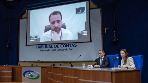 TCE-MS lança plataforma de Inteligência Artificial Generativa e guia de uso ético da tecnologia A meeting at the Tribunal de Contas in Mato Grosso do Sul, featuring a speaker on a video call and two individuals at a table.